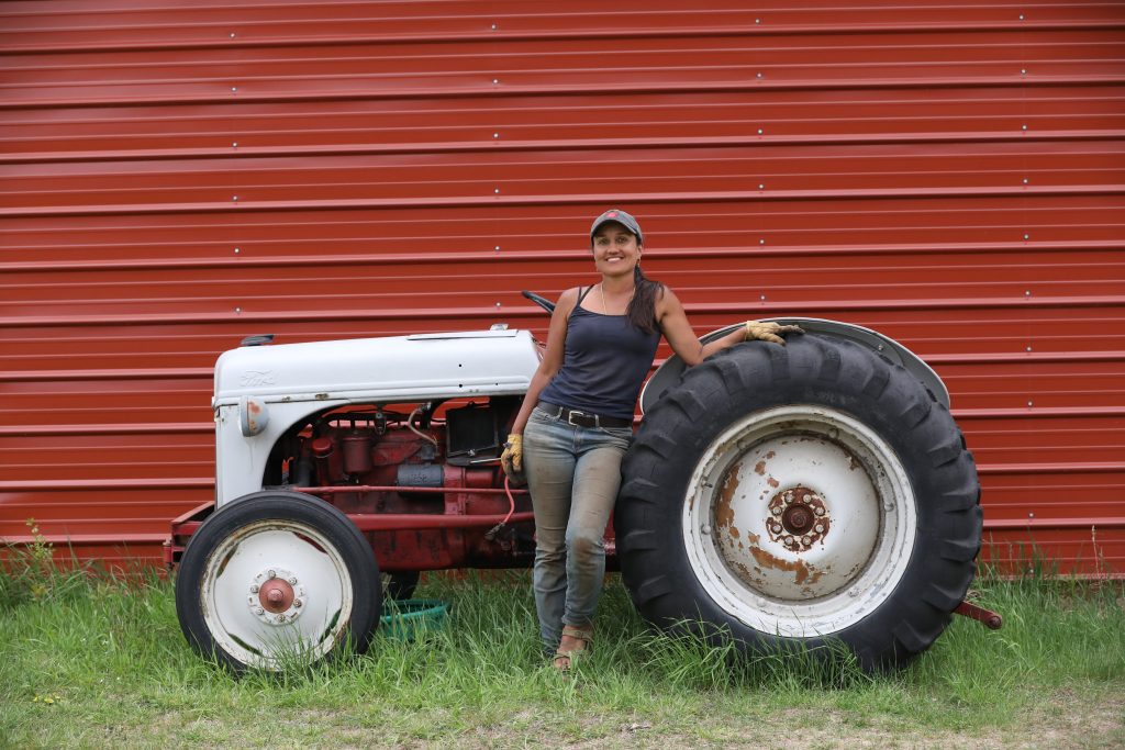 Andal Sundaramurthy stands in front of her tractor with her left arm and the left side of her body resting on an old tractor. Andal and the tractor are in front of a red garage.